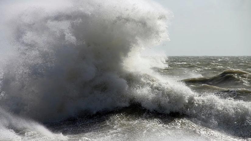 A powerful ocean wave crashing with force, creating a large spray of white foam against a stormy sea backdrop.