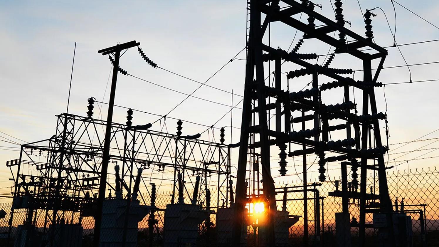 Silhouette of an electrical substation with power lines and transformers against a sunset sky.