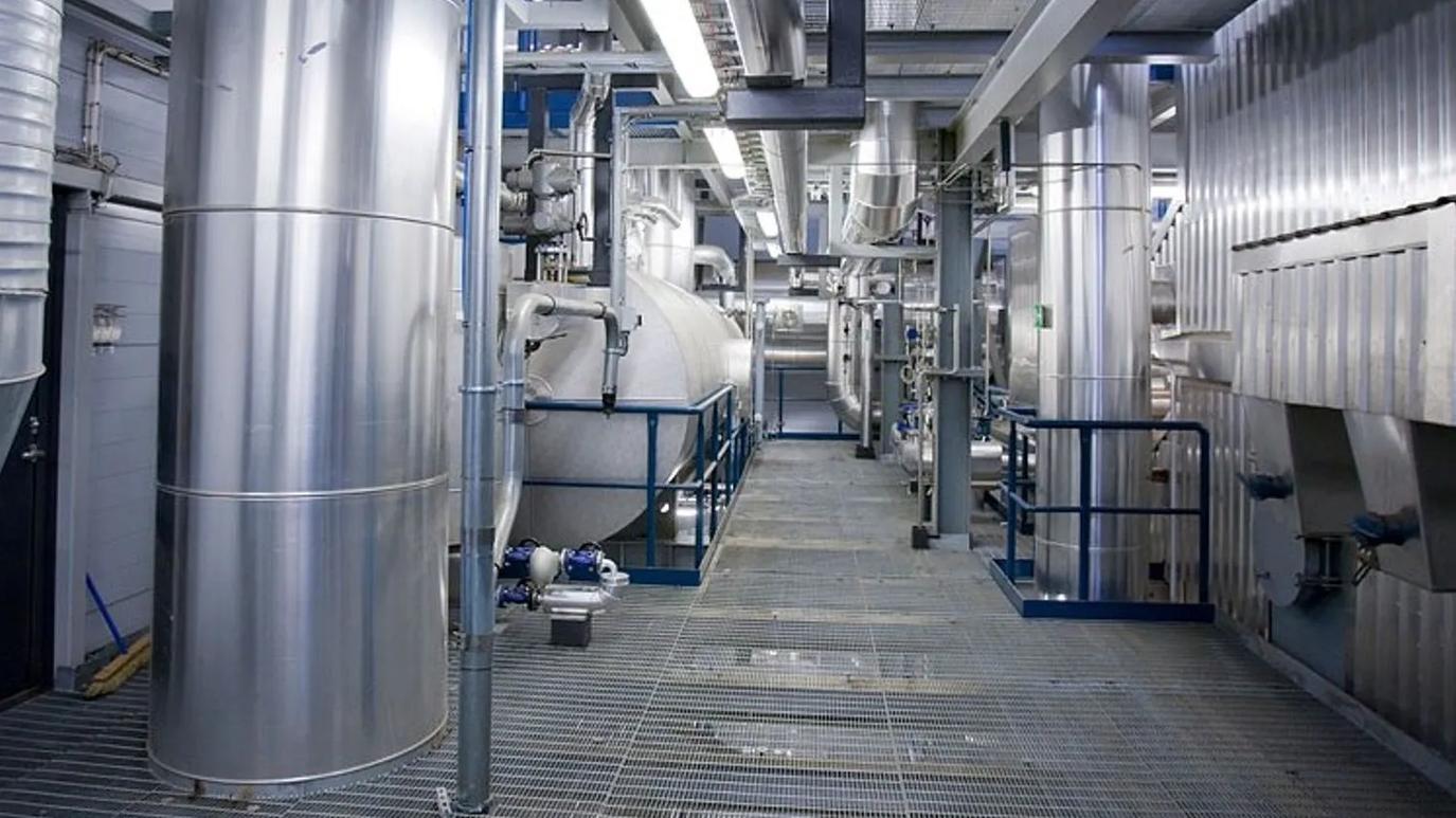 A modern industrial facility interior with large stainless steel tanks and pipes, featuring a grid metal walkway and overhead lighting.