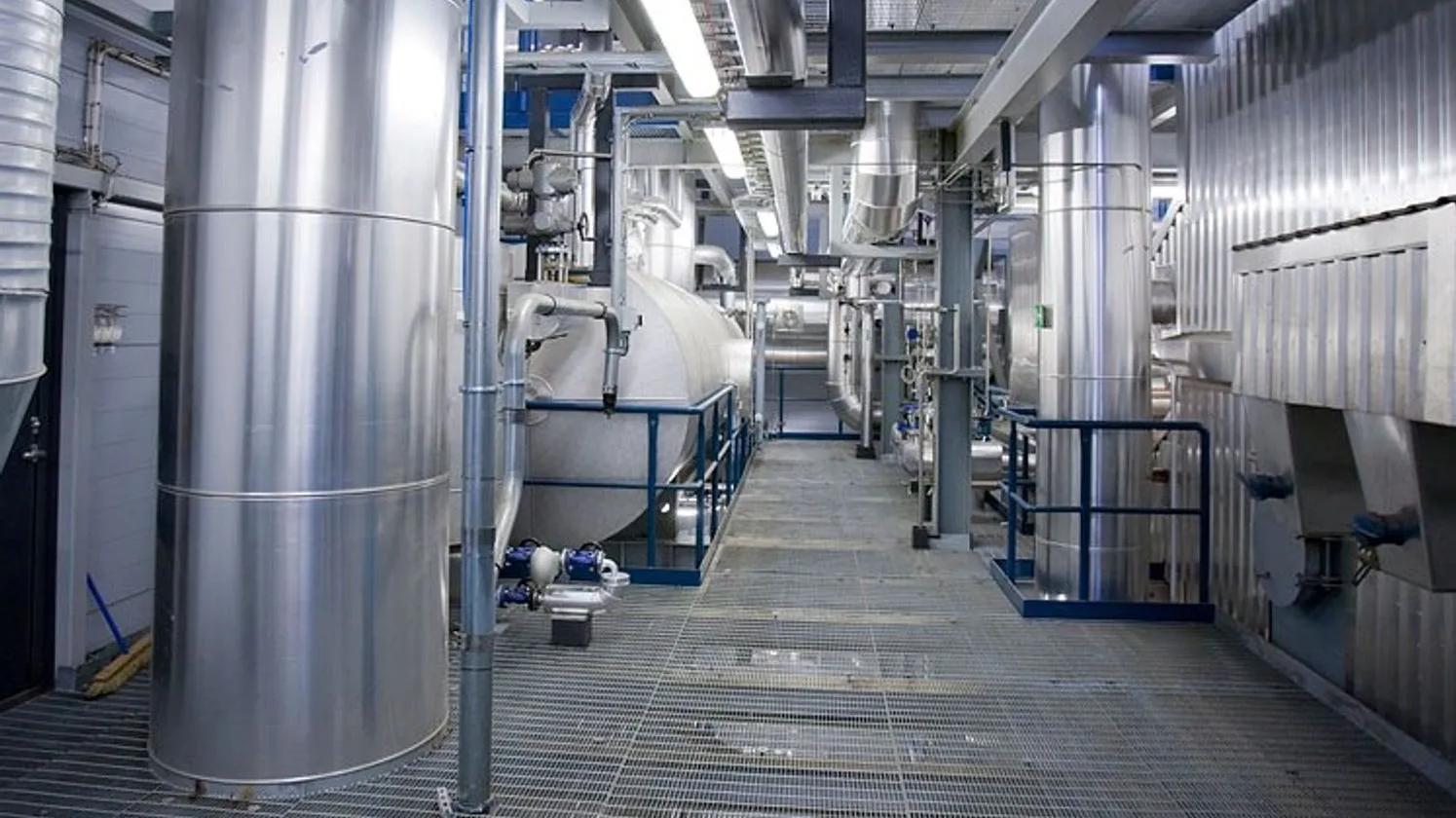 A modern industrial facility interior with large stainless steel tanks and pipes, featuring a grid metal walkway and overhead lighting.