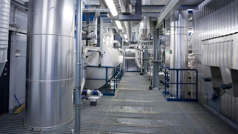 A modern industrial facility interior with large stainless steel tanks and pipes, featuring a grid metal walkway and overhead lighting.