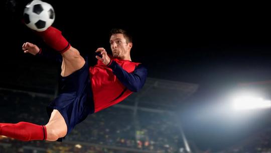 A soccer player in a red and blue uniform performing a mid-air kick with a soccer ball, set against a brightly lit stadium at night.