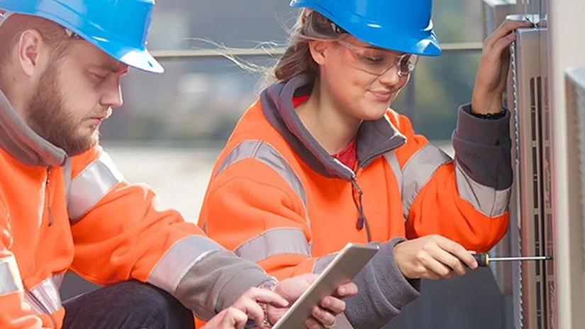 Two construction workers in orange safety jackets and blue helmets, one using a tablet and the other using a screwdriver on equipment.