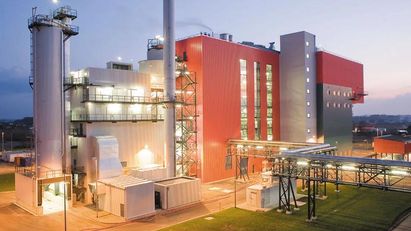 A modern industrial facility with large metal structures and a tall chimney, illuminated at dusk, showcasing a combination of red and silver architectural elements.