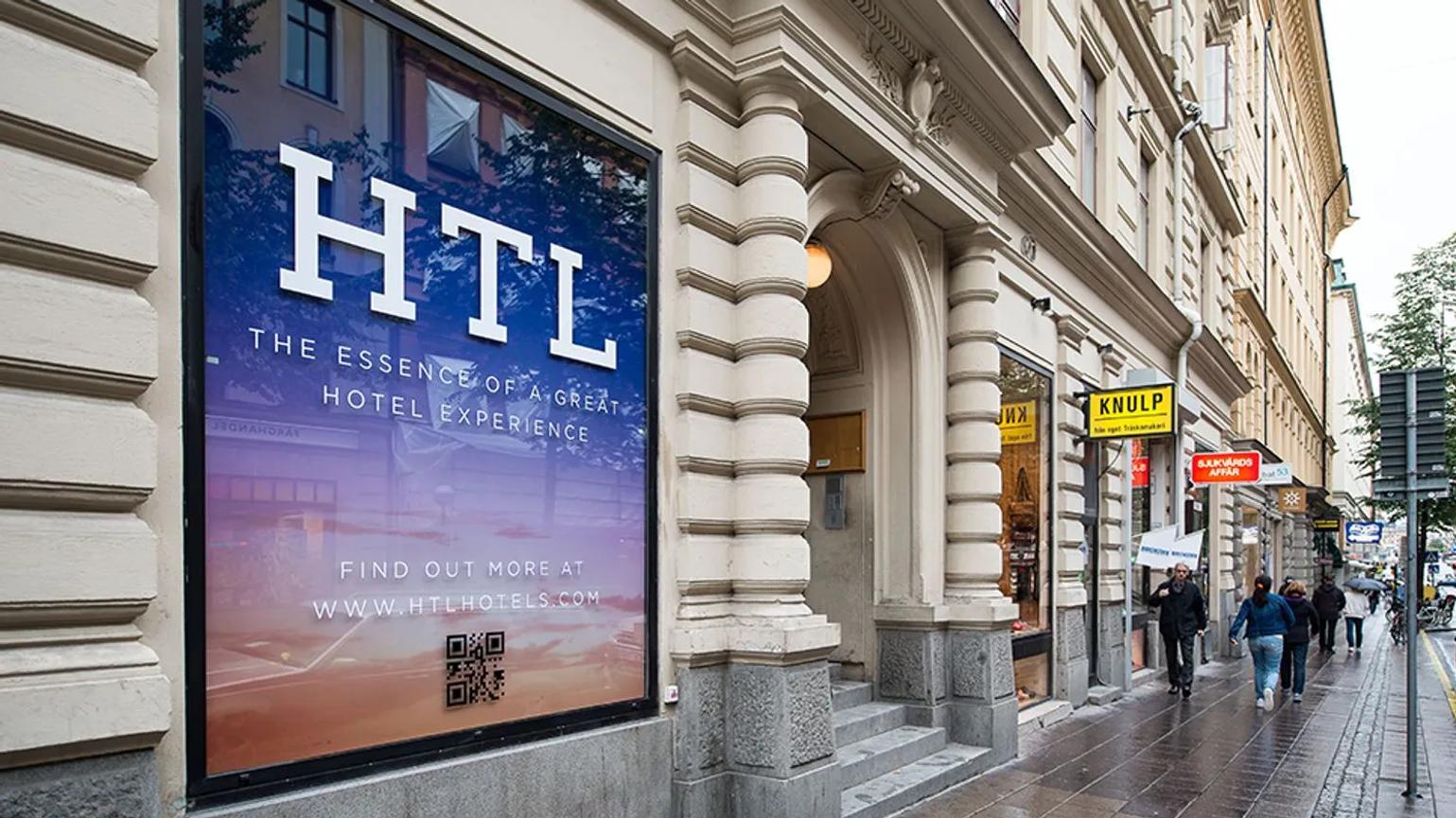 A street view of a building with a large sign displaying "HTL" and the tagline "The Essence of a Great Hotel Experience." The sign includes a QR code and a website link, with pedestrians walking along the sidewalk.