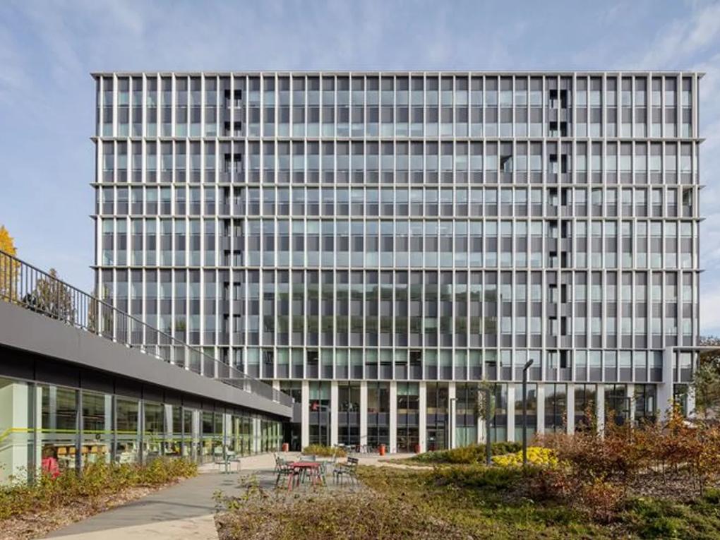 A modern office building with a grid-like facade, featuring large windows and a landscaped outdoor area with tables and chairs.