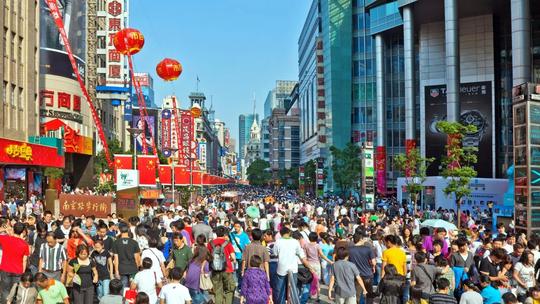 A bustling city street filled with a large crowd of people, surrounded by tall buildings adorned with colorful signs and banners, under a clear blue sky.