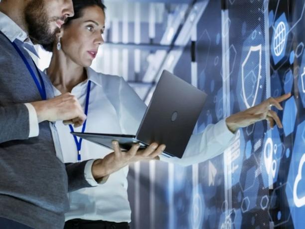 Two professionals in an office setting, one holding a laptop, are interacting with a large digital display.