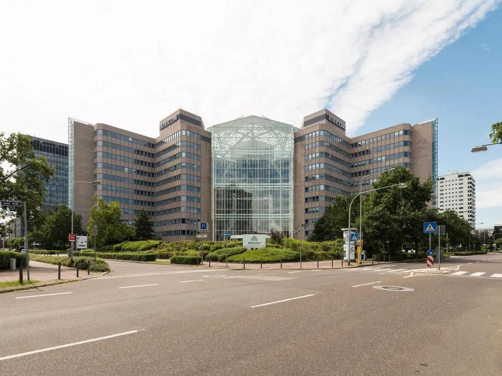 A large office building labeled "Atricom" with a glass atrium in the center, surrounded by greenery and a road in the foreground.