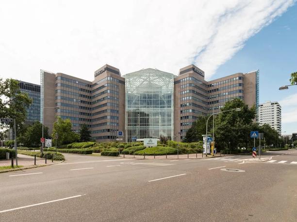 A large office building labeled "Atricom" with a glass atrium in the center, surrounded by greenery and a road in the foreground.