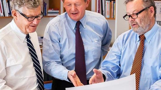Three men in business attire reviewing a large document together in an office with bookshelves in the background.