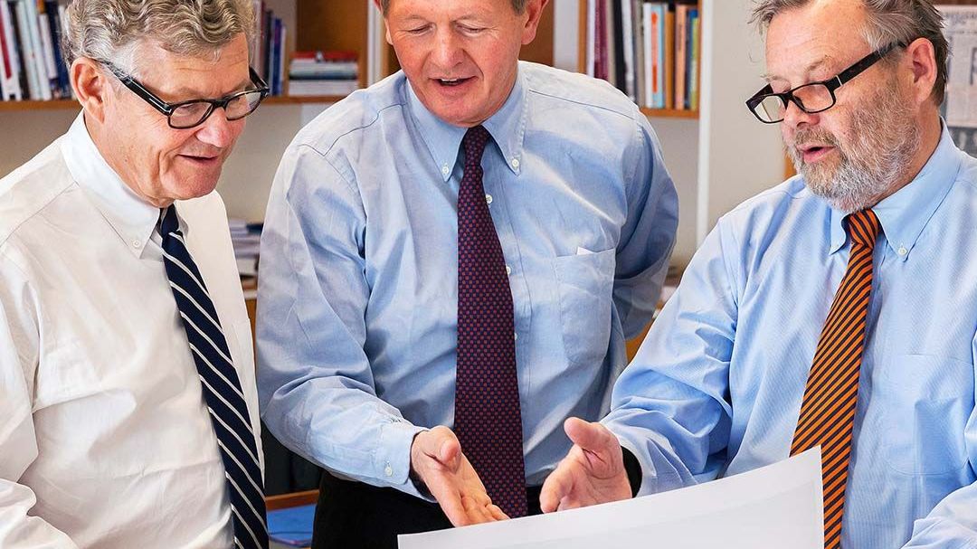 Three men in business attire reviewing a large document together in an office with bookshelves in the background.