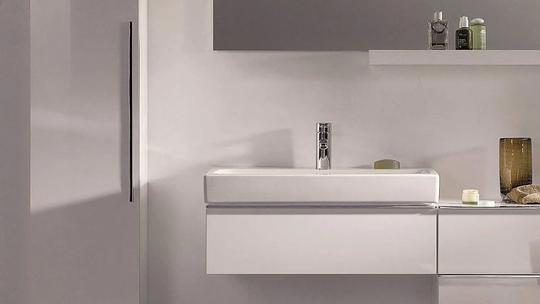 A modern bathroom sink with a sleek design, featuring a chrome faucet and minimalistic cabinetry. The countertop holds a few toiletries and decorative items.