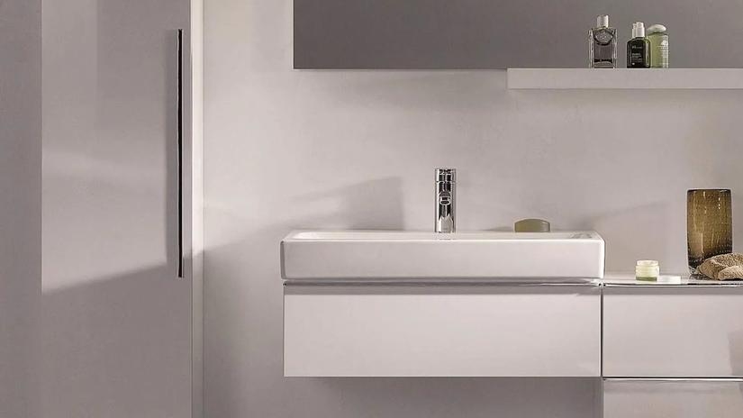 A modern bathroom sink with a sleek design, featuring a chrome faucet and minimalistic cabinetry. The countertop holds a few toiletries and decorative items.