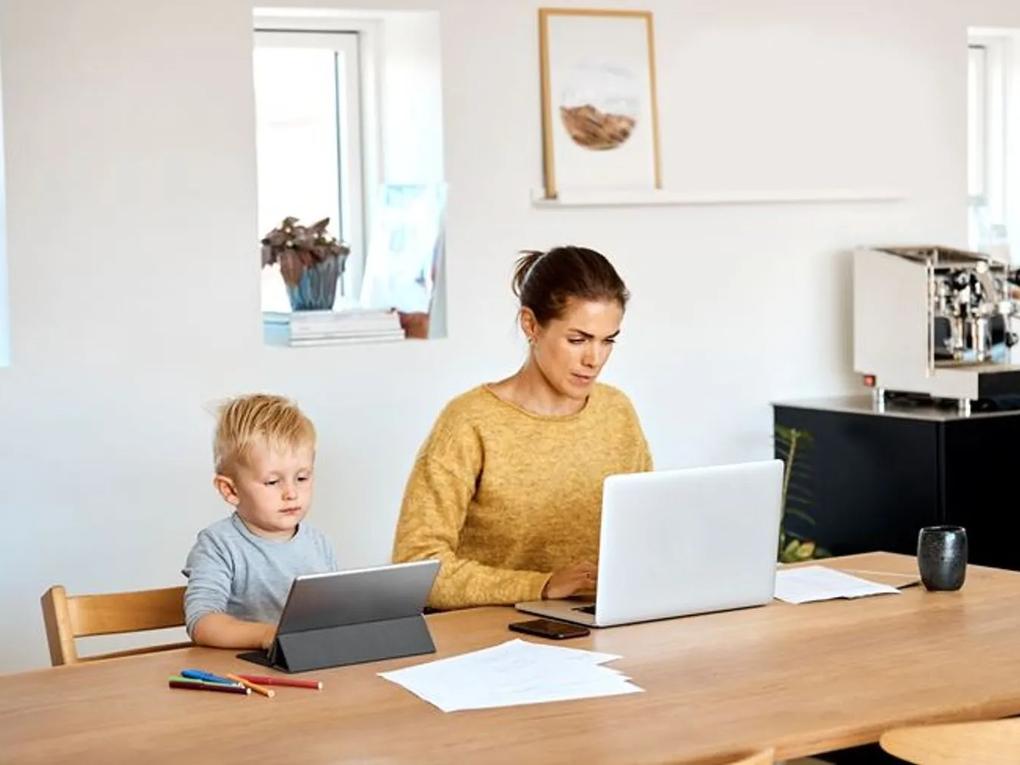 A woman in a yellow sweater working on a laptop at a wooden table, with a young boy beside her using a tablet.