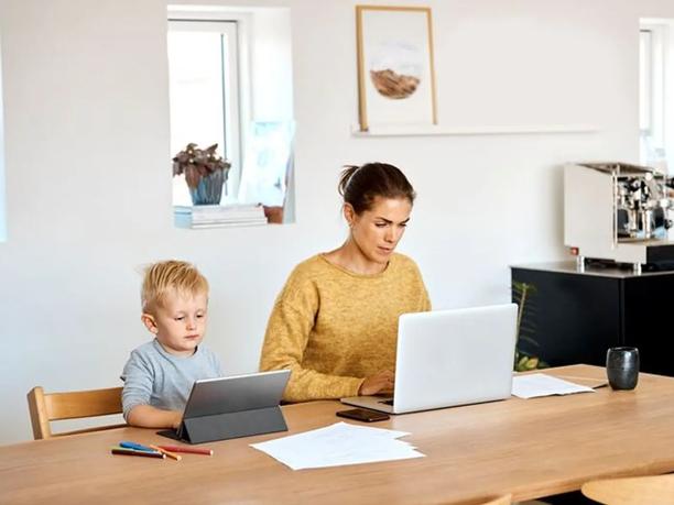 A woman in a yellow sweater working on a laptop at a wooden table, with a young boy beside her using a tablet.