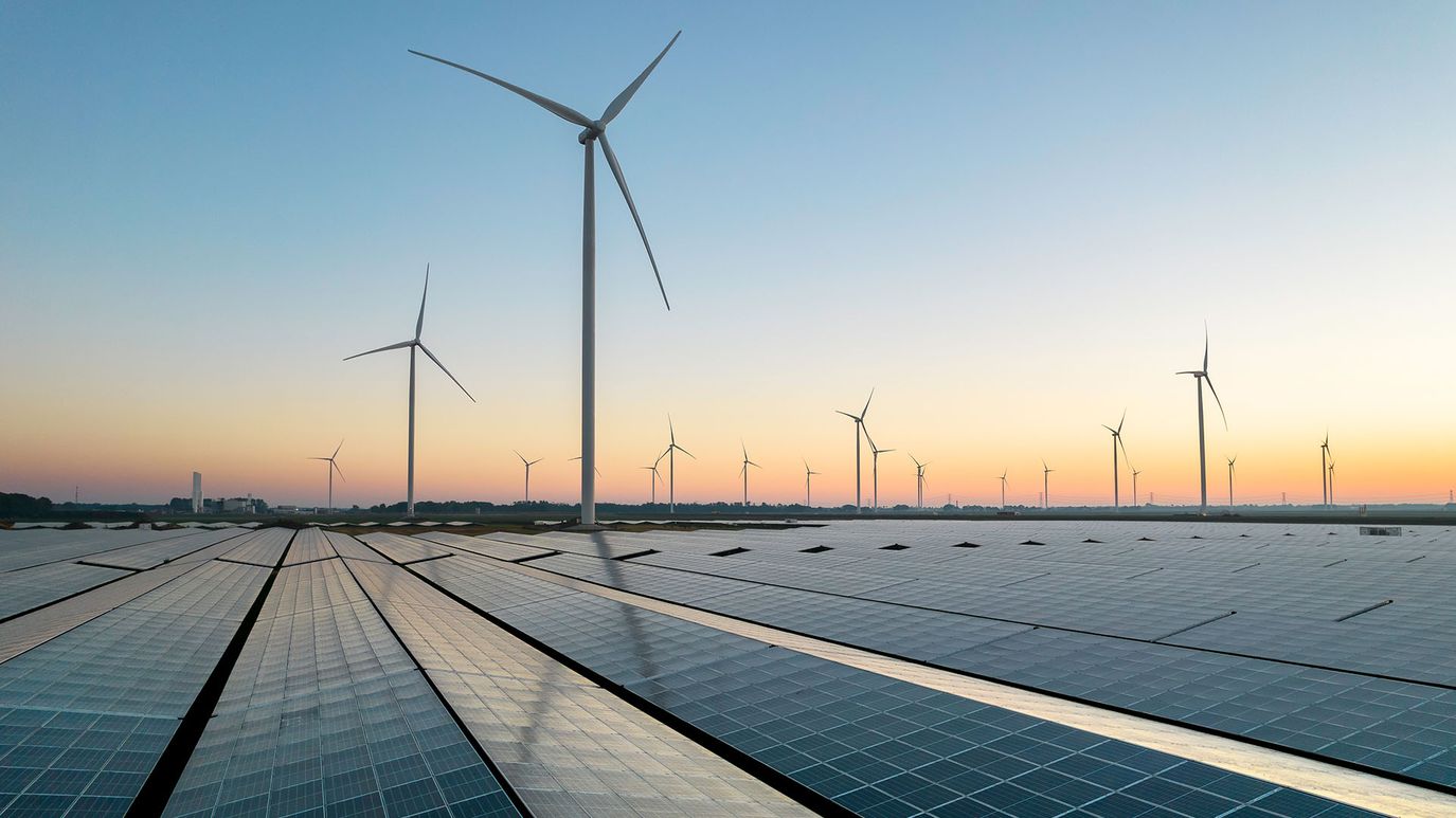 Solar panels in the foreground with multiple wind turbines stretching into the distance at sunset.