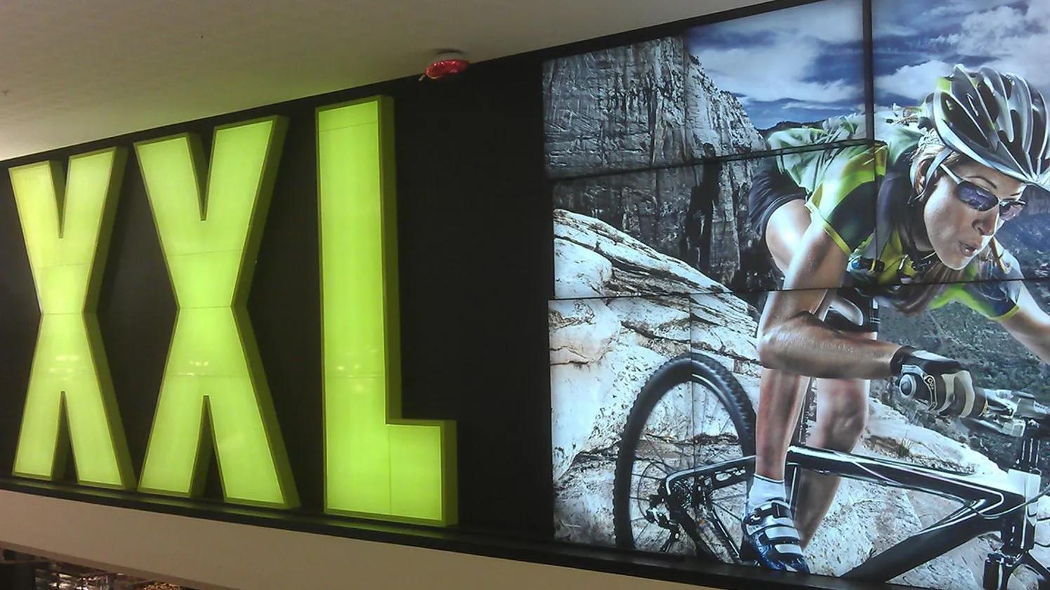 A large illuminated sign with the letters "XXL" next to an image of a cyclist wearing a helmet and sunglasses, riding a mountain bike on a rocky trail.