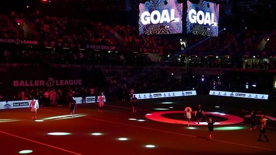 Indoor football match at a Baller League event with players on a dark pitch, colorful spotlights, and large screens displaying "GOAL."
