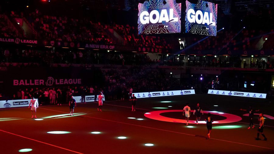 Indoor football match at a Baller League event with players on a dark pitch, colorful spotlights, and large screens displaying "GOAL."