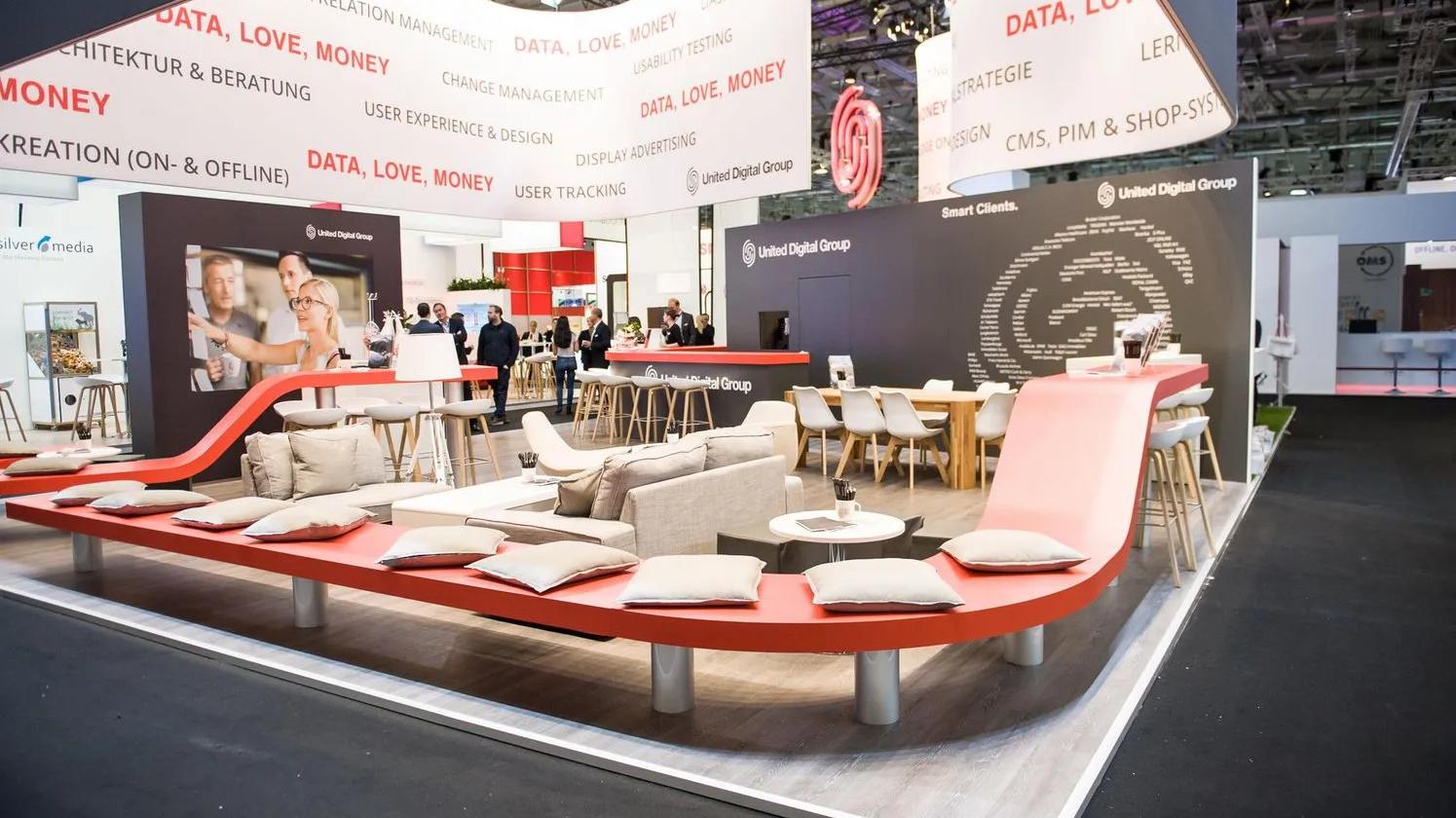 A modern exhibition booth for United Digital Group, featuring sleek red and white seating areas with cushions, tables, and digital displays. The booth is adorned with phrases like "DATA, LOVE, MONEY" and includes a large logo on the wall.