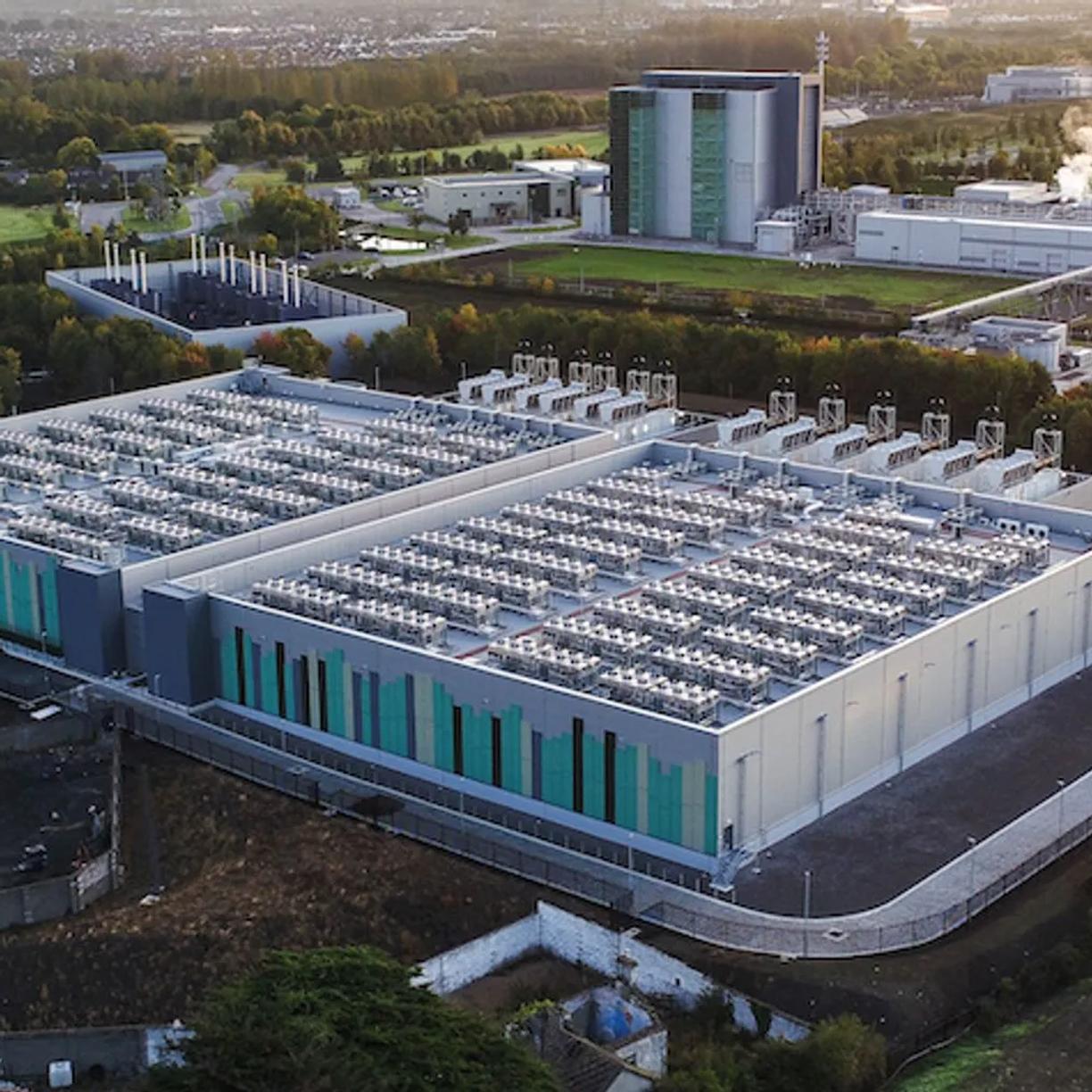 Aerial view of a large data center with multiple cooling units on the roof, surrounded by greenery and other industrial buildings in the background.