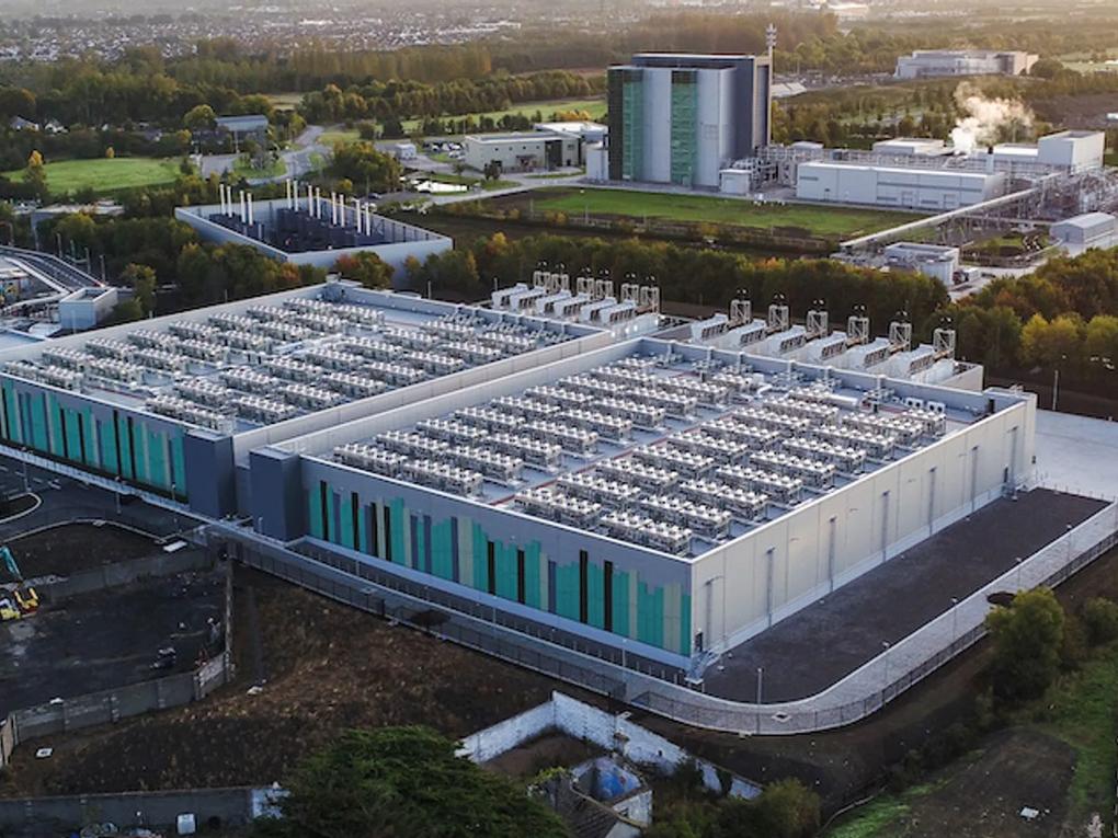 Aerial view of a large data center with multiple cooling units on the roof, surrounded by greenery and other industrial buildings in the background.