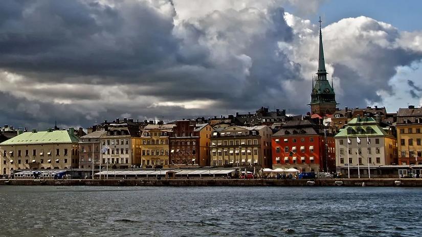 A waterfront view of the old town in Stockholm, featuring colorful historic buildings and a prominent church spire under a dramatic cloudy sky.