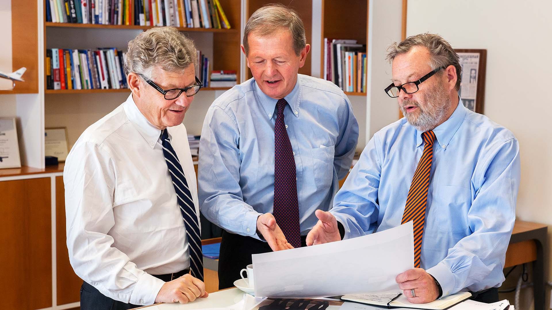 Three men in business attire reviewing a large document together in an office with bookshelves in the background.