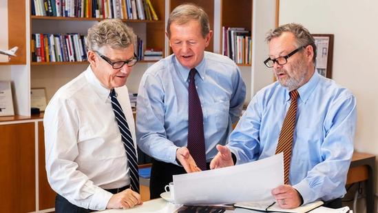 Three men in business attire reviewing a large document together in an office with bookshelves in the background.