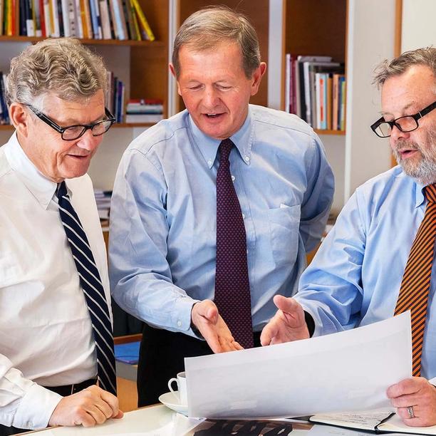 Three men in business attire reviewing a large document together in an office with bookshelves in the background.