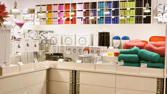 A retail display featuring colorful home goods, including wall clocks, lamps, and decorative cushions. The shelves are neatly organized with vibrant storage boxes and various household items under hanging lights.