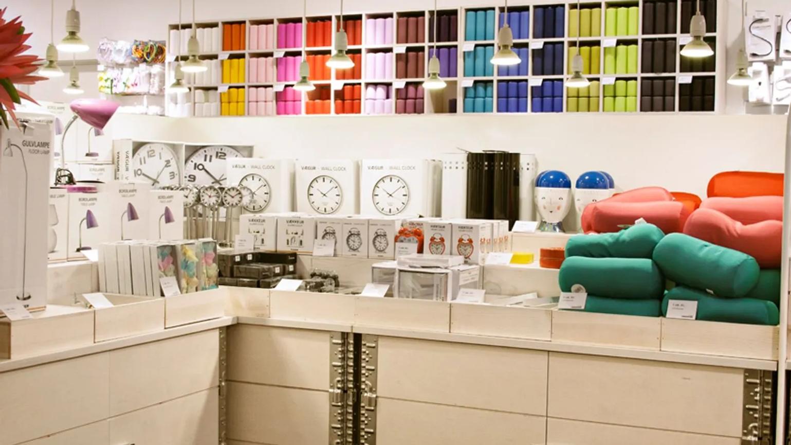 A retail display featuring colorful home goods, including wall clocks, lamps, and decorative cushions. The shelves are neatly organized with vibrant storage boxes and various household items under hanging lights.