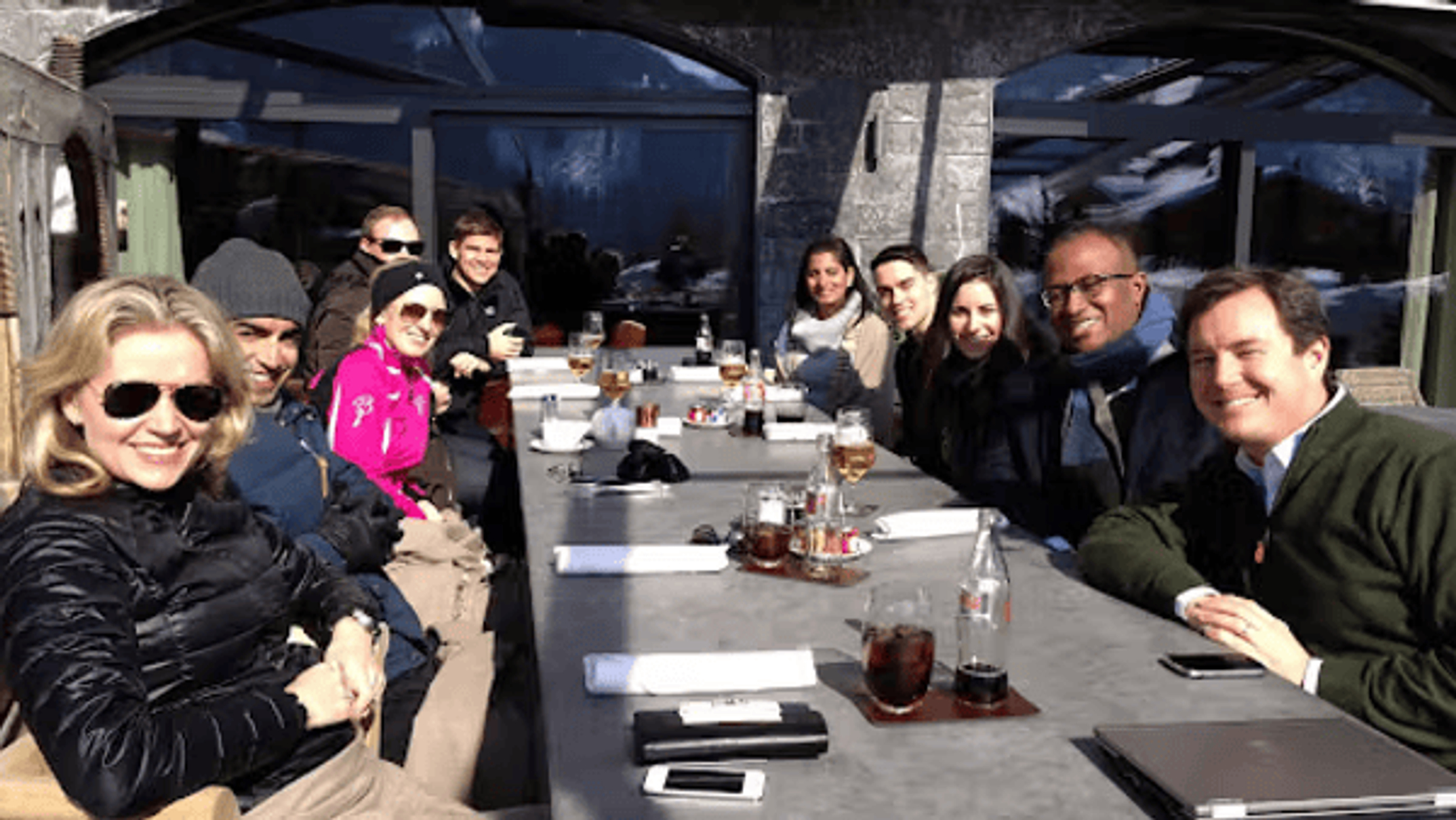 A group of people sitting around a long outdoor table, enjoying drinks and smiling in a sunny setting.