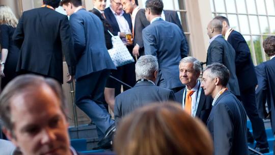 A group of business professionals in suits, some wearing lanyards, gathered outside a building, engaging in conversation and networking.