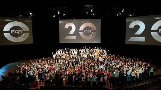 A large group of people celebrating in an auditorium with two screens displaying the EQT logo and the number 20, indicating an anniversary or milestone event.'