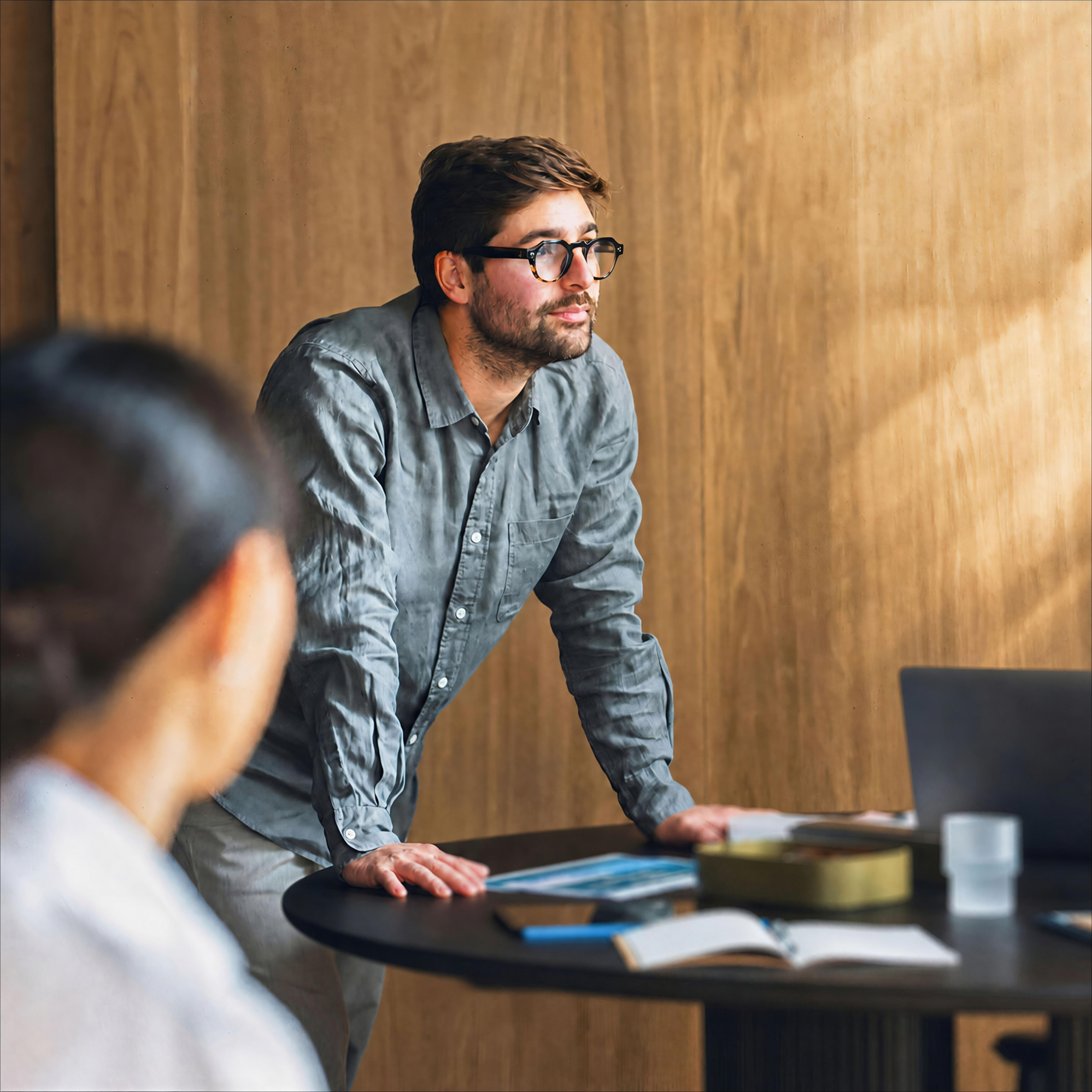 A man with glasses and a beard standing at a round meeting table, leaning forward on his hands while looking toward the side during a discussion.