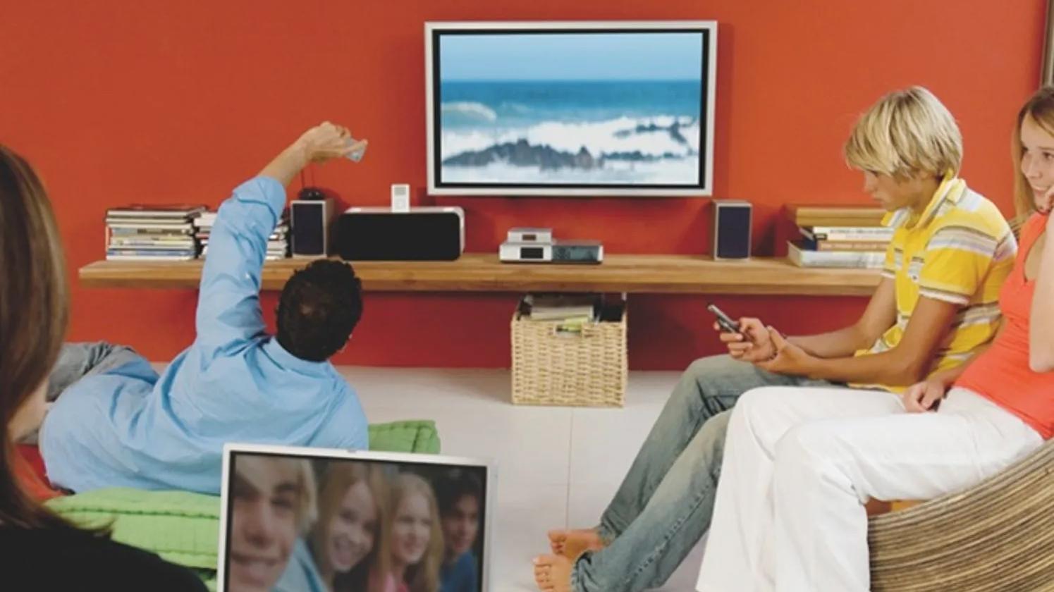A group of people sitting in a living room watching a television, with one person using a remote control and another using a smartphone. The room has a modern design with a red accent wall and a wooden shelf holding electronic devices.