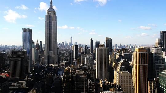 A panoramic view of New York City featuring the Empire State Building prominently in the foreground, surrounded by other skyscrapers under a clear blue sky.