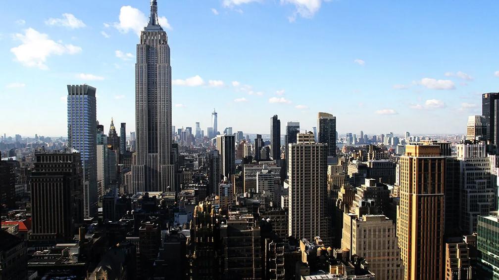 A panoramic view of New York City featuring the Empire State Building prominently in the foreground, surrounded by other skyscrapers under a clear blue sky.