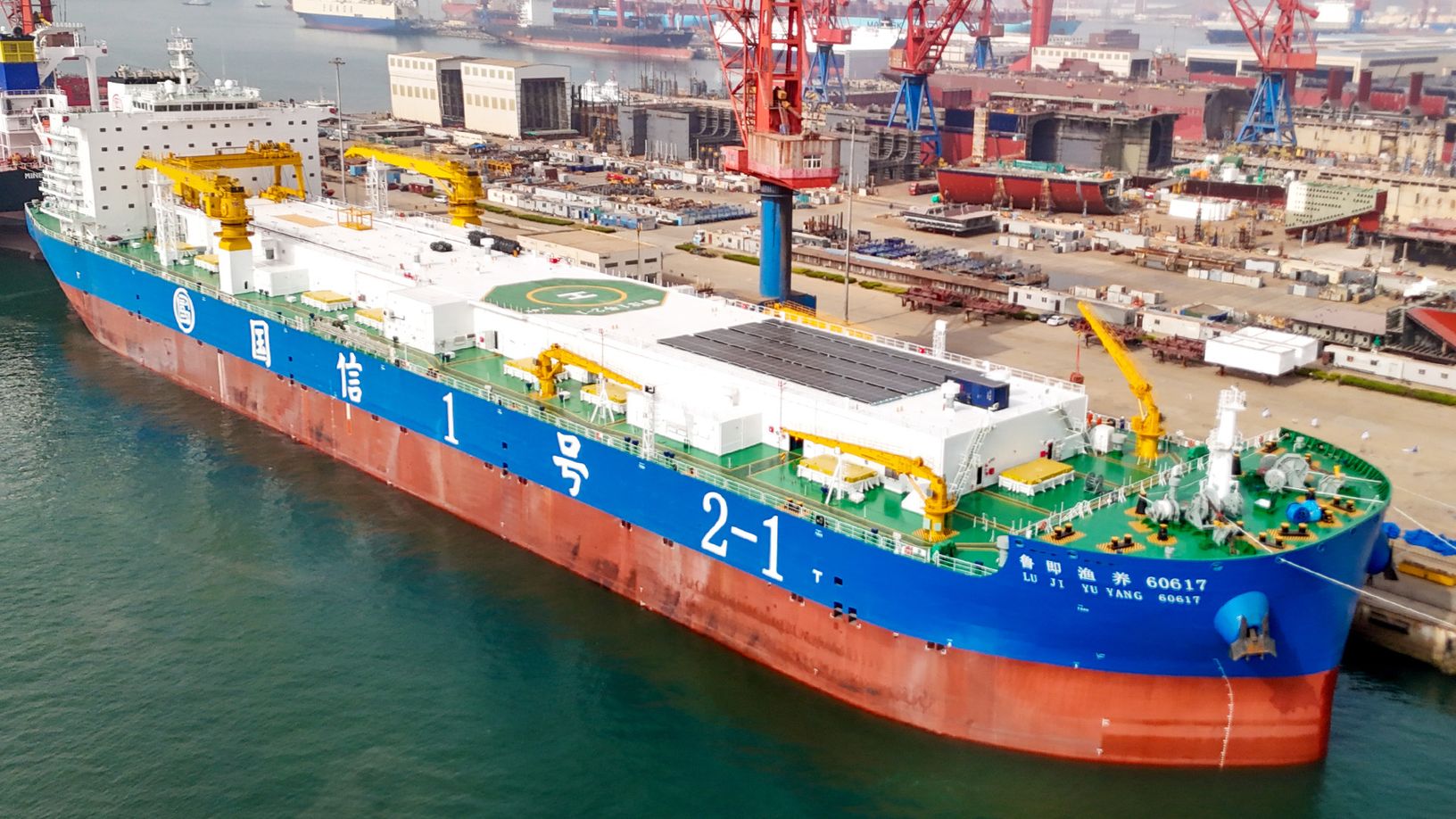 A large blue and white industrial ship with cranes and a helipad is docked at a busy shipyard, surrounded by other vessels and port equipment.