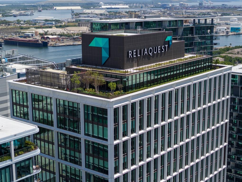 Aerial view of a modern high-rise office building with a rooftop garden and the large sign 'ReliaQuest' on the top. The building is surrounded by other glass buildings and overlooks a waterfront industrial area with ships and docks in the background.