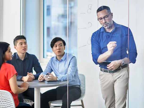 A man in a blue shirt writing on a glass board, while three colleagues sit at a table, watching attentively.