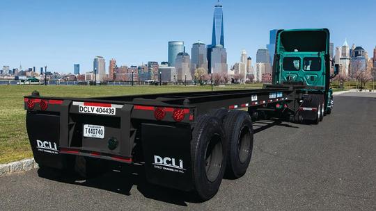 A green truck with a flatbed trailer parked on a road, with the New York City skyline in the background.