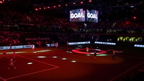 Indoor football match at a Baller League event with players on a dark pitch, colorful spotlights, and large screens displaying "GOAL."