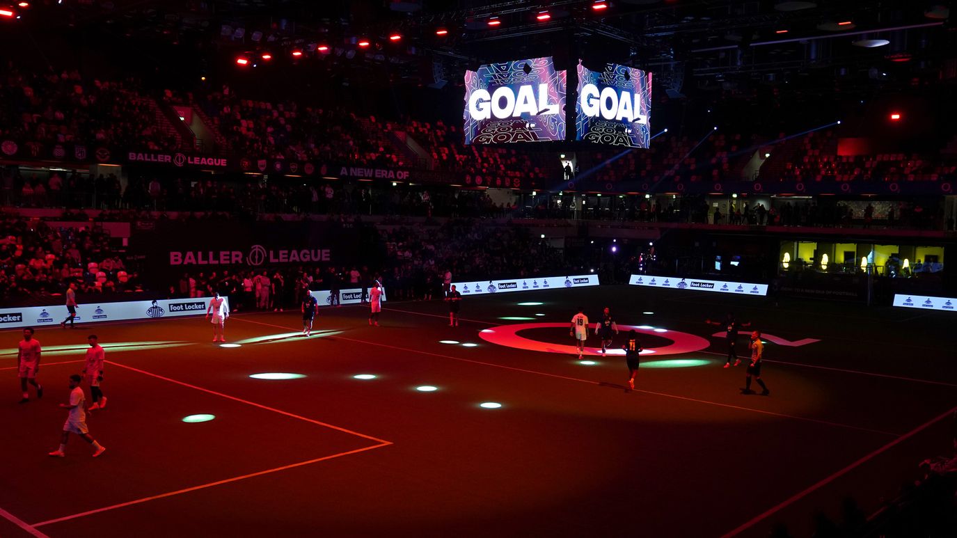 Indoor football match at a Baller League event with players on a dark pitch, colorful spotlights, and large screens displaying "GOAL."
