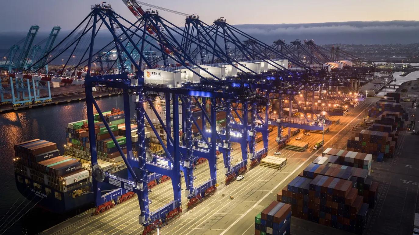 Aerial view of a busy shipping port at dusk, featuring large blue cranes labeled "Fenix" and stacks of colorful shipping containers.