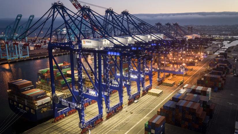 Aerial view of a busy shipping port at dusk, featuring large blue cranes labeled "Fenix" and stacks of colorful shipping containers.