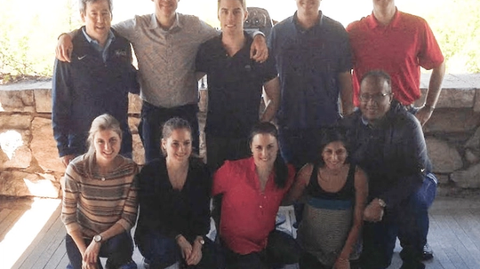 A group of nine people posing together, with some standing and others kneeling, in an outdoor setting.
