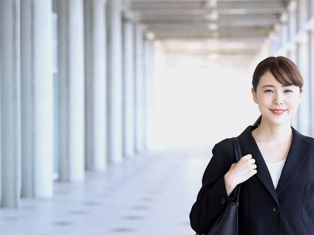 A smiling woman in a black blazer holding a bag, standing in a bright corridor.